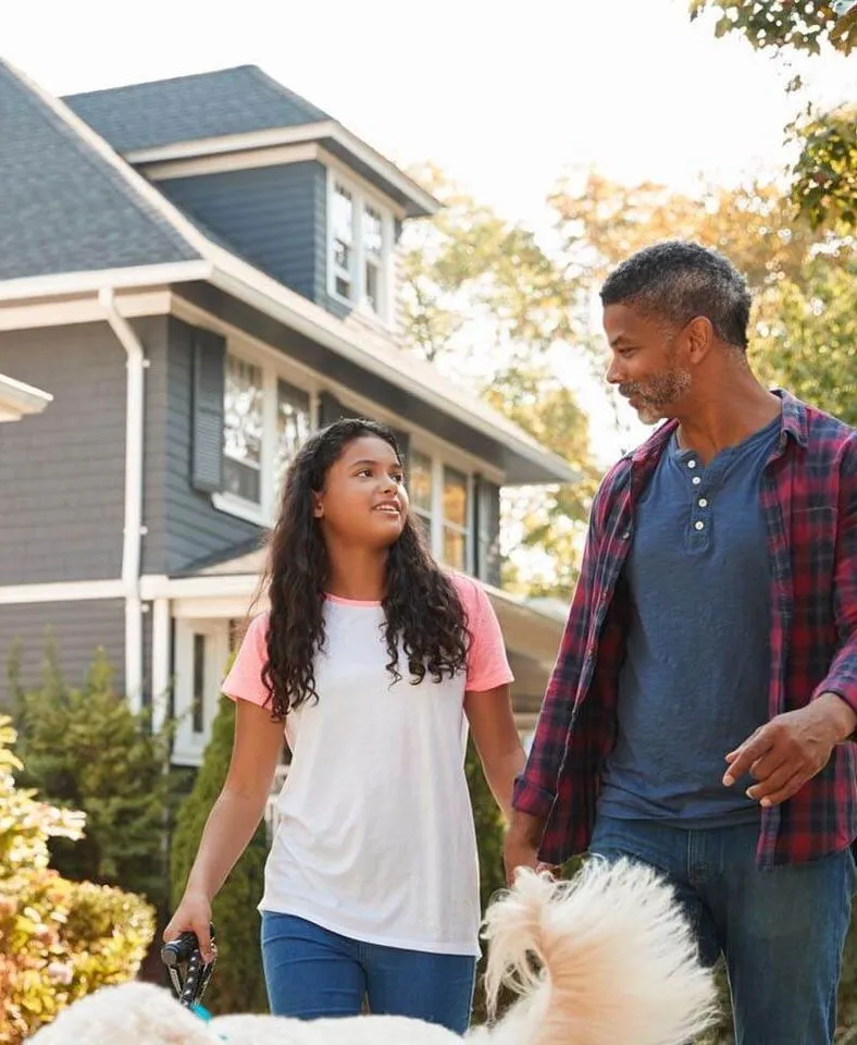 Father and daughter walking a dog in front of a house.